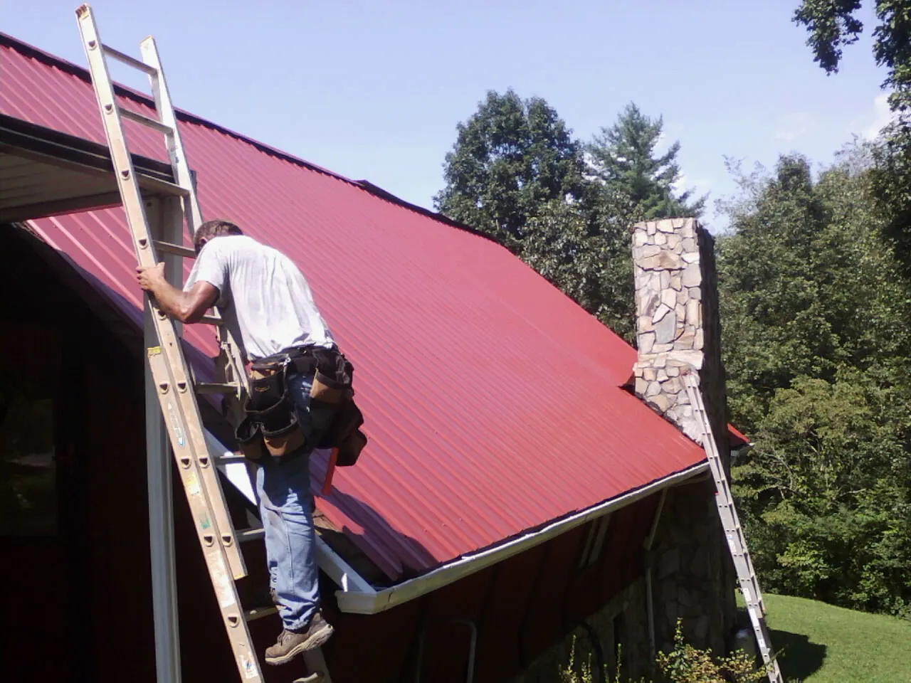 Roofer on a ladder beside a red metal roof and stone chimney — Soffit & Fascia Repair by Roofing Brothers in Zanesfield